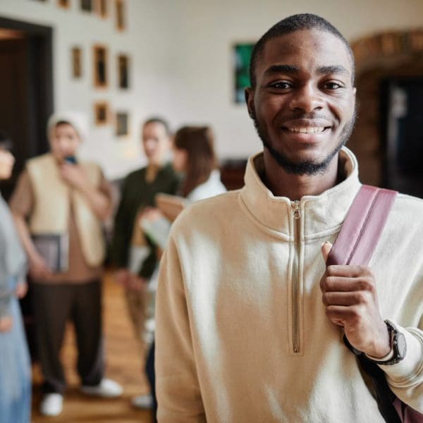 A young Black student smiles at the camera while holding his backpack strap, with a group of blurred classmates chatting in the background.