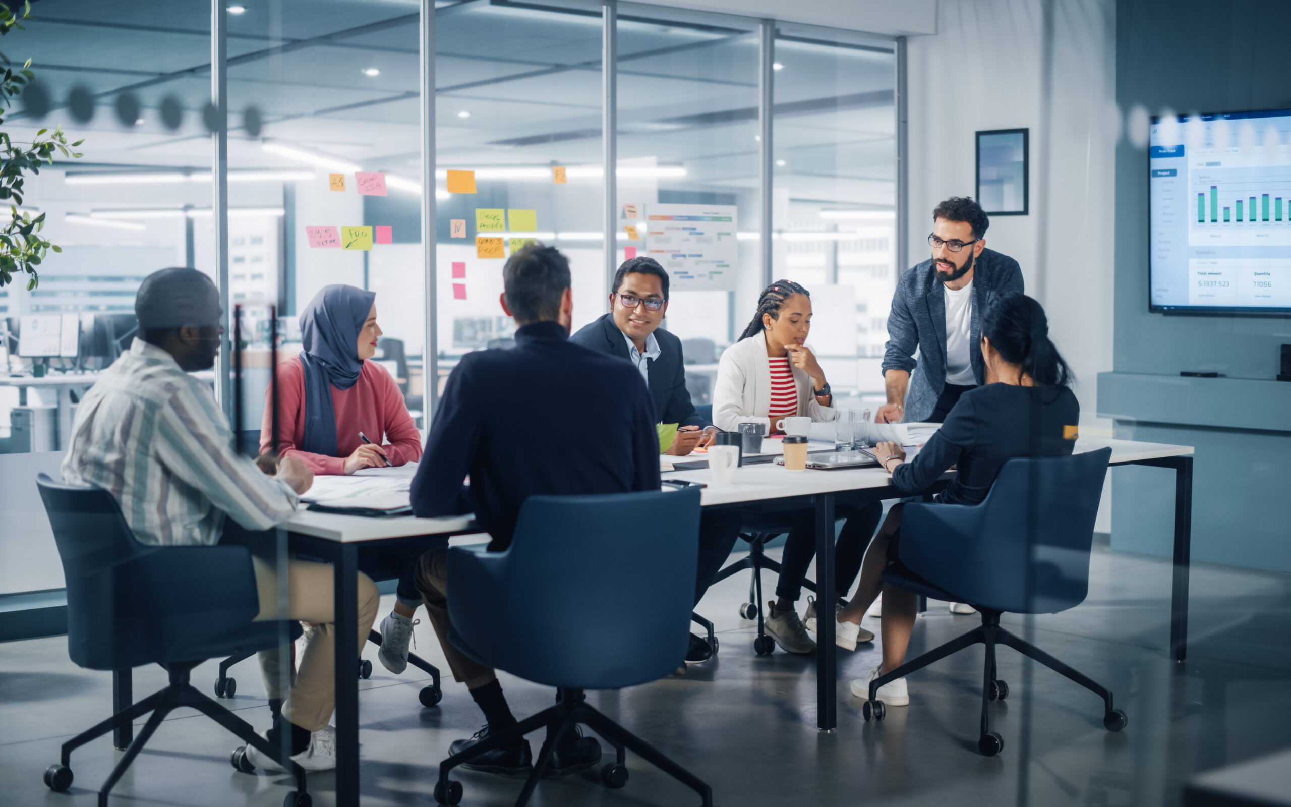 A diverse corporate team sits around a conference table in a modern glass office, listening to a standing colleague present data from a wall screen.