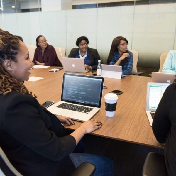 A diverse group of professionals sits around a large wooden conference table with laptops open, smiling and engaged in a lively office meeting.