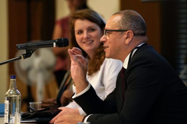 A man in a suit and glasses gestures while speaking into a microphone, while a woman sits next to him and smiles attentively.