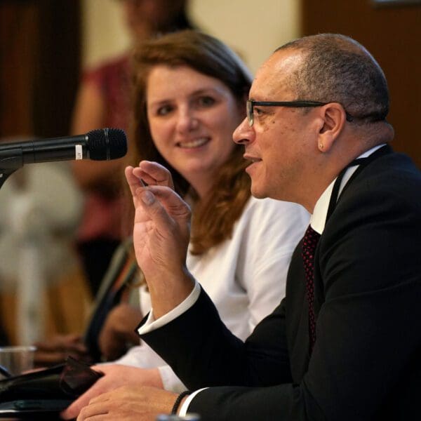 A man in a suit and glasses gestures while speaking into a microphone, while a woman sits next to him and smiles attentively.