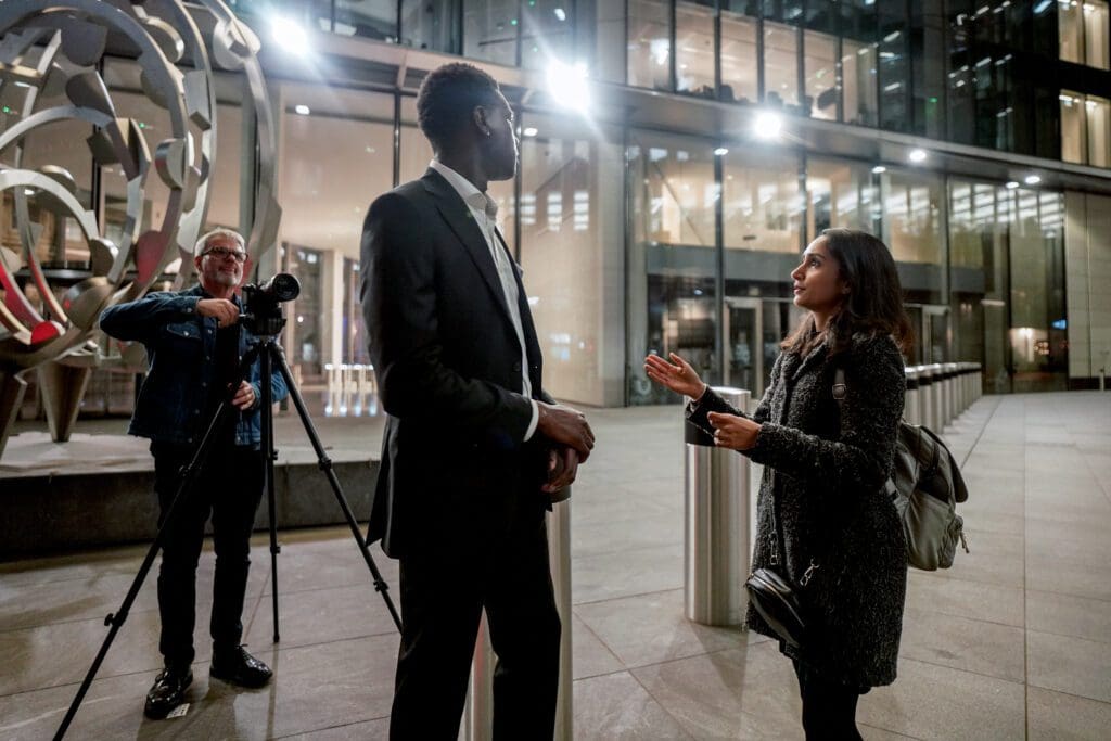 Two people facing each other in an interview, outside of a shiny glass building in the city. A cameraman points his lens on a tripod at the pair.