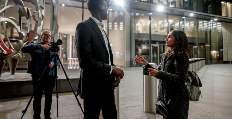 Two people facing each other in an interview, outside of a shiny glass building in the city. A cameraman points his lens on a tripod at the pair.