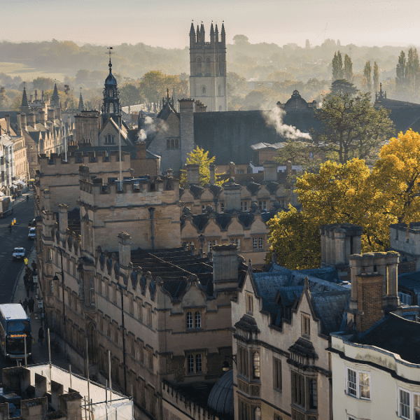 An elevated view of Oxford's historic skyline features stone collegiate buildings, a curving street with a double-decker bus, autumn trees, and distant towers under morning mist.