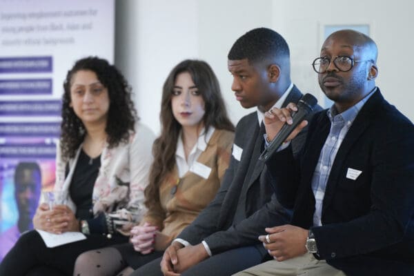 A diverse panel of four professionals sits at a table during an event. One man speaks into a microphone while his colleagues listen attentively.