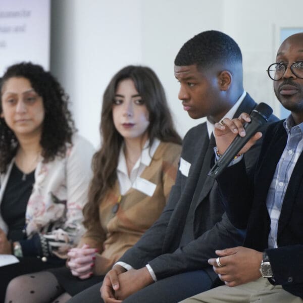 A diverse panel of four professionals sits at a table during an event. One man speaks into a microphone while his colleagues listen attentively.