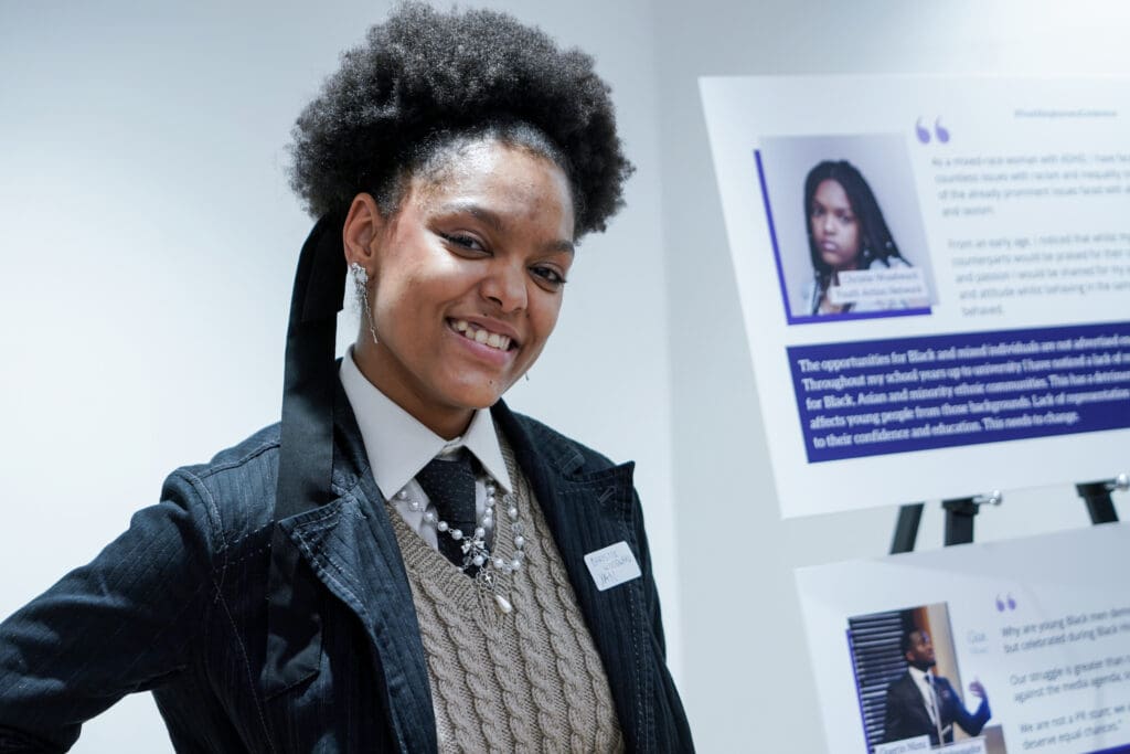 A smiling young Black woman with an afro hairstyle stands in front of display boards. She wears a denim jacket, cable-knit vest, and a tie with necklaces