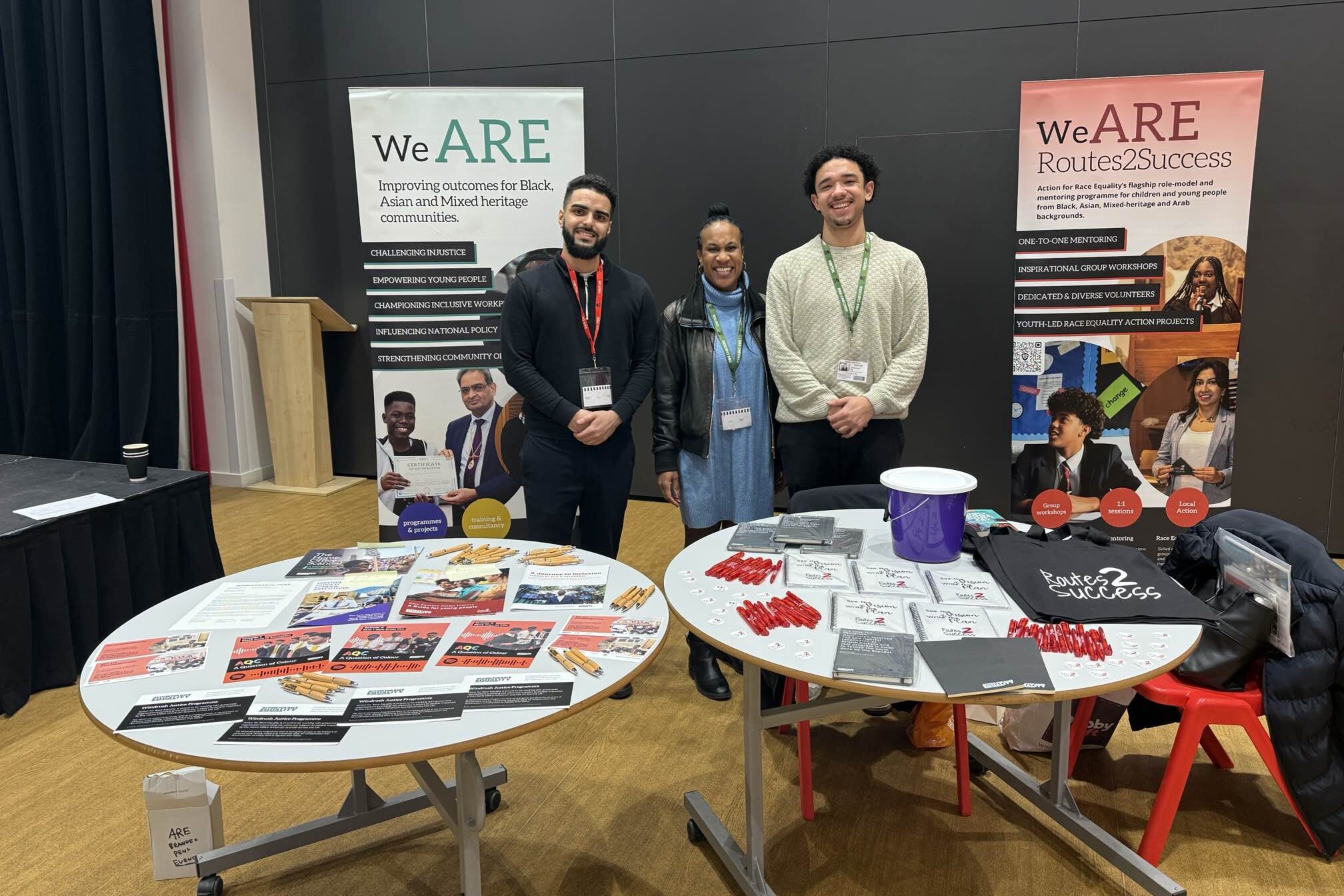 three people standing at an Action for Race Equality event with tables displaying brochures and banners behind them.