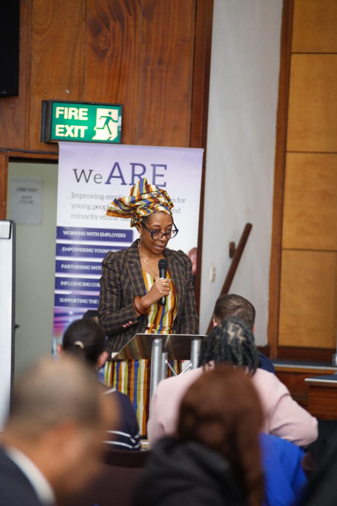 A woman wearing a patterned headwrap and blazer speaking at a podium during the Northern Cities Youth Employment Conference, addressing an audience.