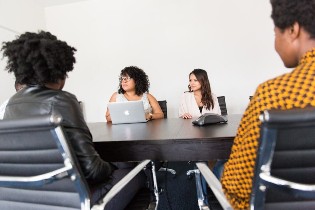 Four women of differing ethnic groups, sit around a boardroom table. Their gaze is towards someone out of shot.