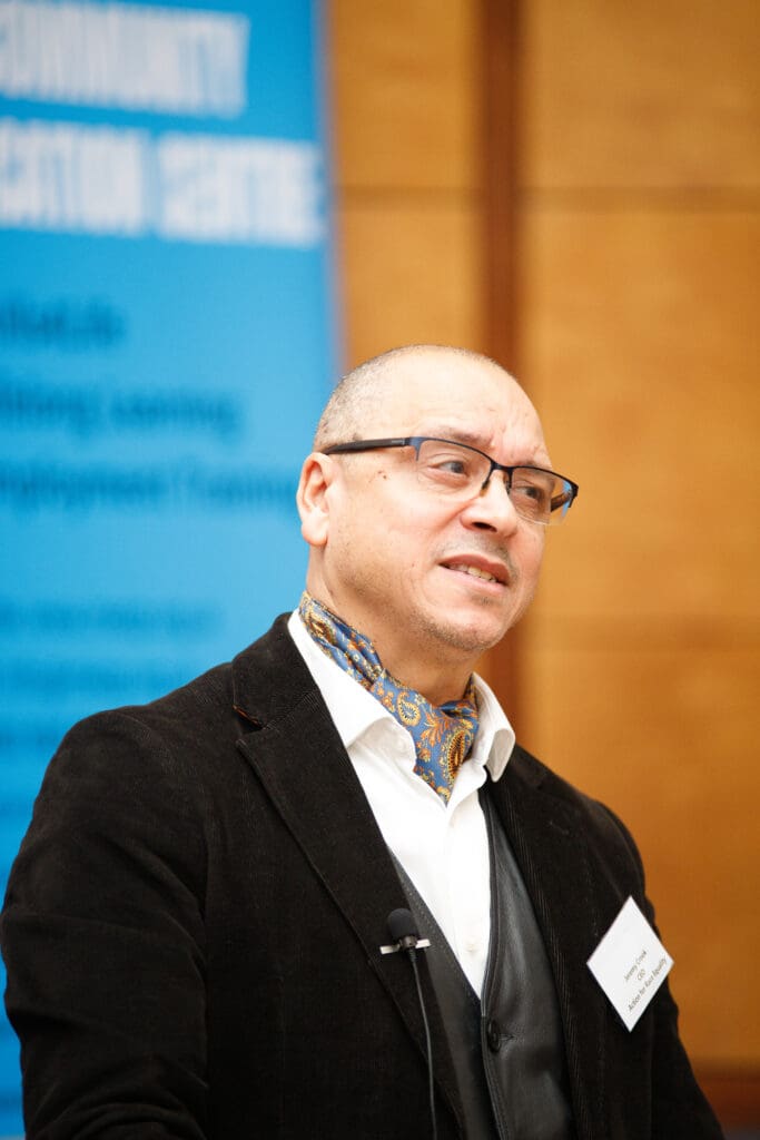 A man wearing glasses, a black blazer, and a patterned scarf speaking at the Northern Cities Youth Employment Conference, standing in front of a blue backdrop.