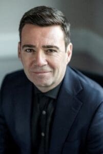 A headshot of a smiling man with short dark hair, wearing a dark navy blazer over a black button-down shirt.
