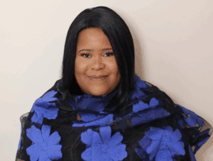 A headshot of a smiling Black woman with straight dark hair, draped in a black fabric wrap featuring large, vibrant blue floral patterns against a plain, light background.