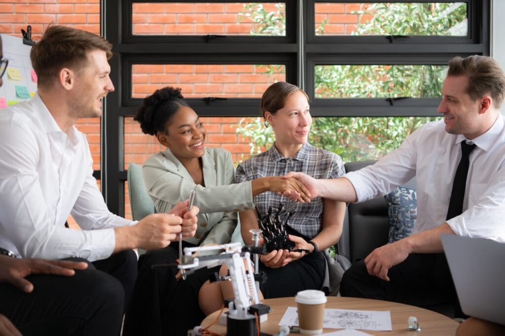 A diverse group of professionals smiles as a Black woman and a white man shake hands across a table. Small robotic arms sit on the table between them.