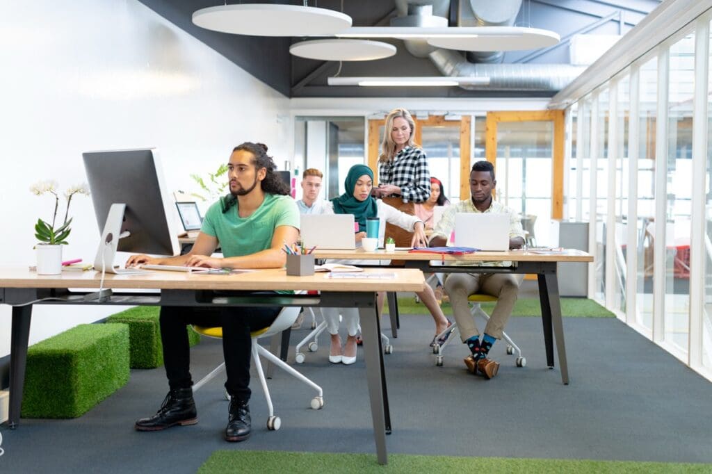 A diverse group of professionals work on computers in a bright, modern open-plan office. One woman stands while others sit at long desks with laptops and a monitor.
