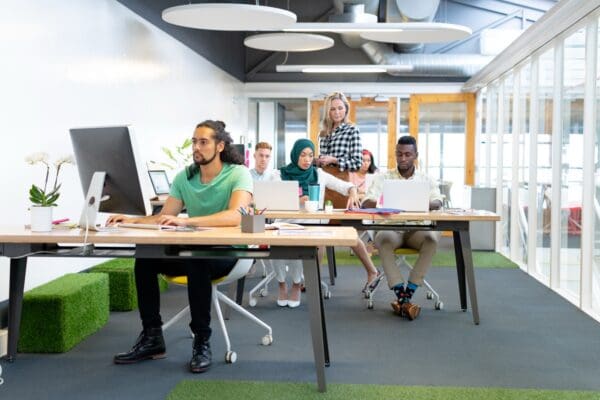 A diverse group of professionals work on computers in a bright, modern open-plan office. One woman stands while others sit at long desks with laptops and a monitor.