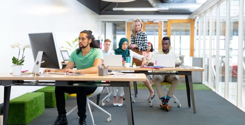 A diverse group of professionals work on computers in a bright, modern open-plan office. One woman stands while others sit at long desks with laptops and a monitor.