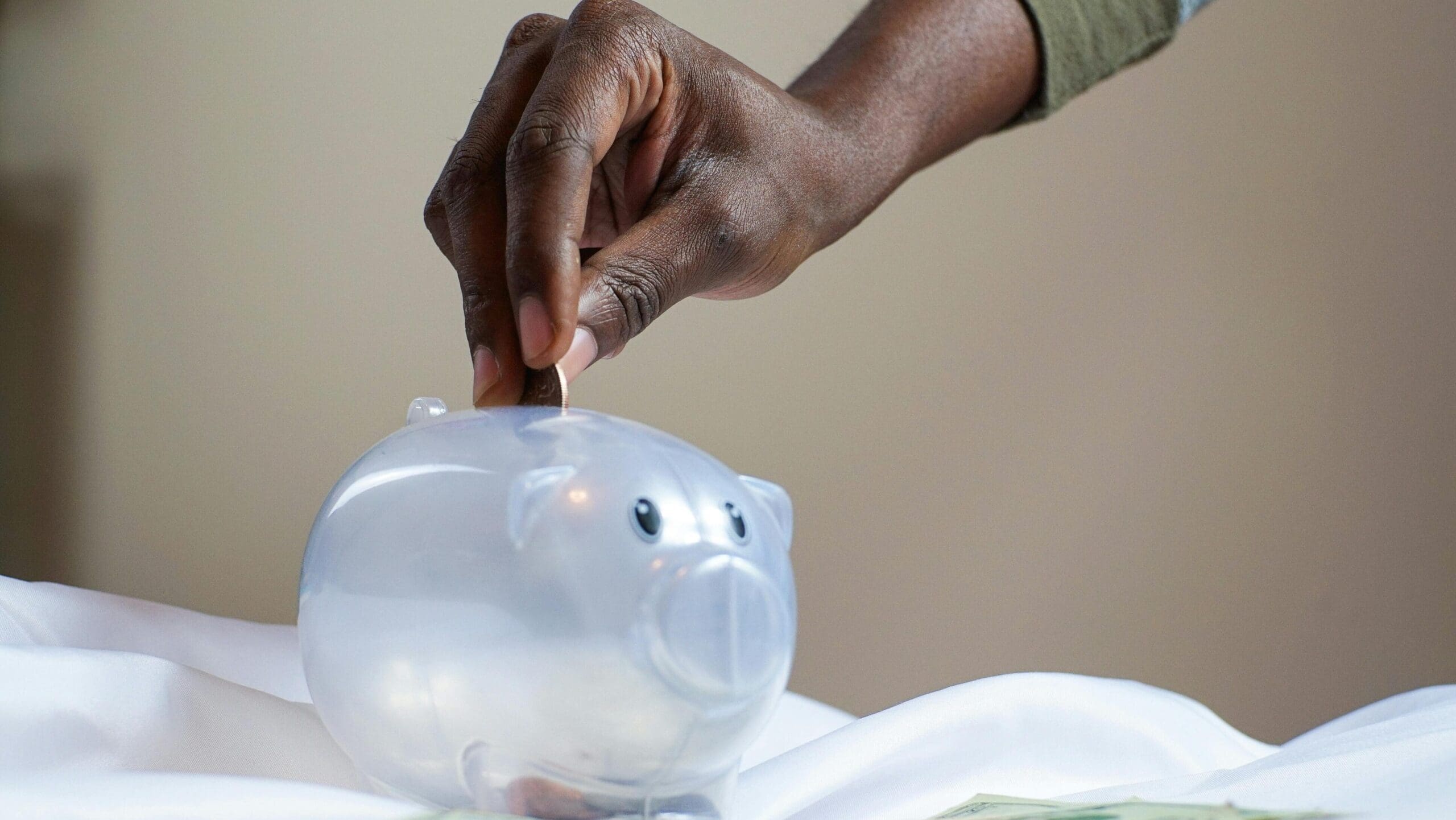 Close-up of a person's hand placing coins into a transparent piggy bank to save money.