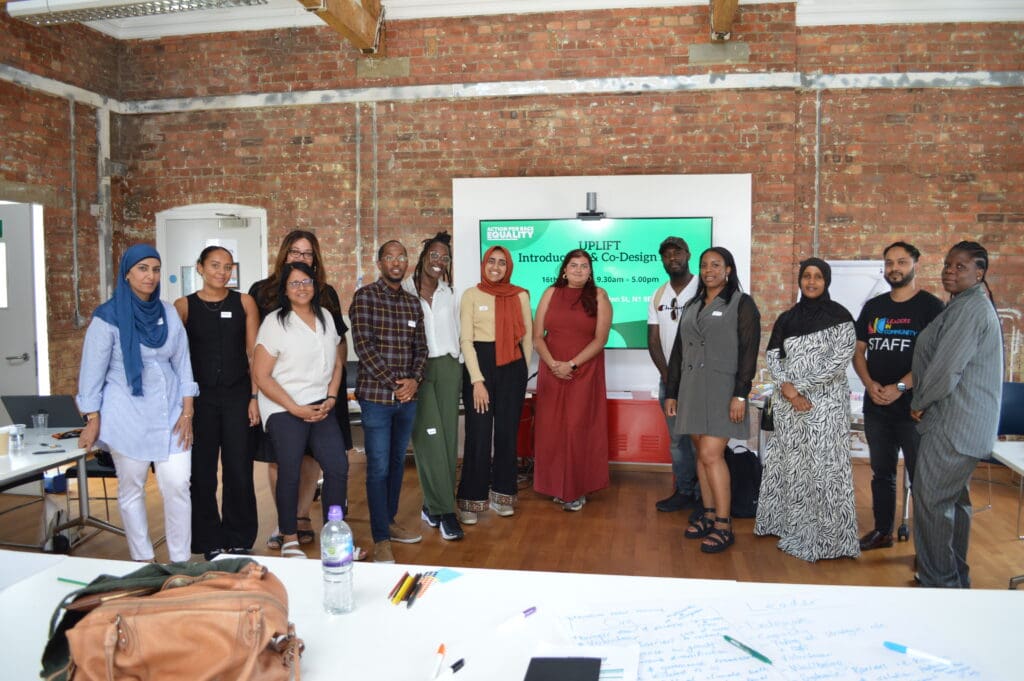 A diverse group of twelve people stand together for a group photo in a room with exposed brick walls. A presentation screen for "UPLIFT" is behind them.