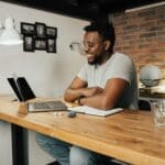 African American man smiling and working remotely from home using a laptop and digital tablet.