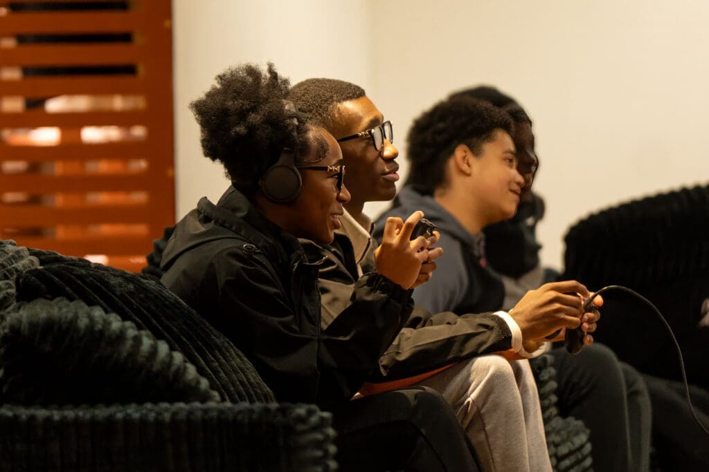Two people sit on a dark, textured sofa, each holding a wired game controller, appearing focused on gameplay. Both wear casual clothing, with one in a black jacket and headphones, and the other in a hoodie. A third person is partially visible in the background. The setting appears indoors with warm lighting and a wooden panel feature on the wall. Faces are blurred for privacy.