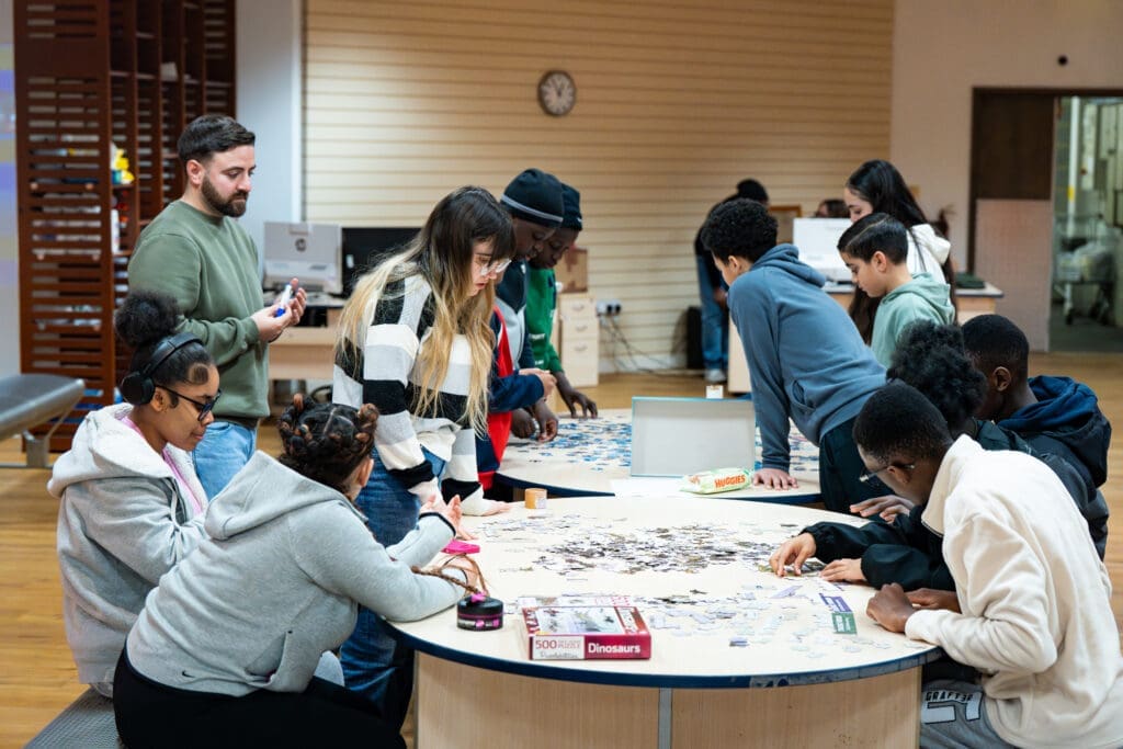 A group of people are gathered around two round tables in a brightly lit indoor space, working together on large jigsaw puzzles. Puzzle pieces are spread across the tables, with some participants seated and others standing. A puzzle box labeled “Dinosaurs” is visible in the foreground. In the background, there are computers, shelves, and a clock on the wall.