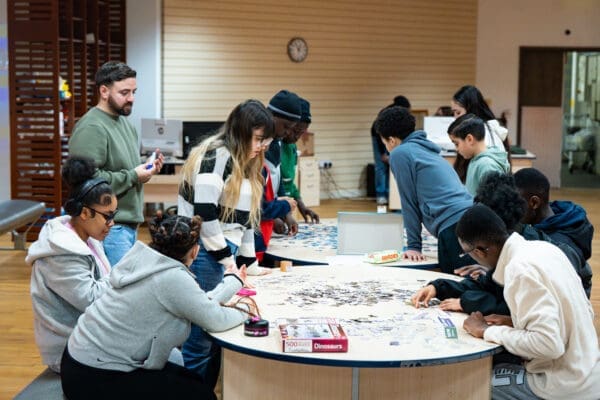 A group of people are gathered around two round tables in a brightly lit indoor space, working together on large jigsaw puzzles. Puzzle pieces are spread across the tables, with some participants seated and others standing. A puzzle box labeled “Dinosaurs” is visible in the foreground. In the background, there are computers, shelves, and a clock on the wall.