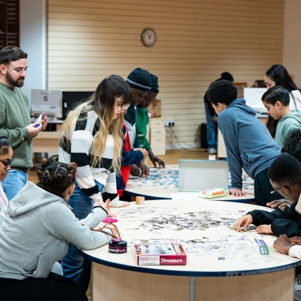 A group of people are gathered around two round tables in a brightly lit indoor space, working together on large jigsaw puzzles. Puzzle pieces are spread across the tables, with some participants seated and others standing. A puzzle box labeled “Dinosaurs” is visible in the foreground. In the background, there are computers, shelves, and a clock on the wall.