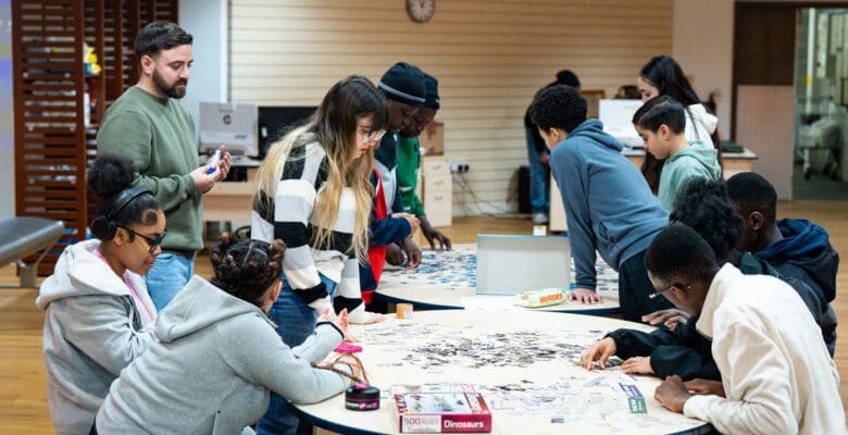 A group of people are gathered around two round tables in a brightly lit indoor space, working together on large jigsaw puzzles. Puzzle pieces are spread across the tables, with some participants seated and others standing. A puzzle box labeled “Dinosaurs” is visible in the foreground. In the background, there are computers, shelves, and a clock on the wall.