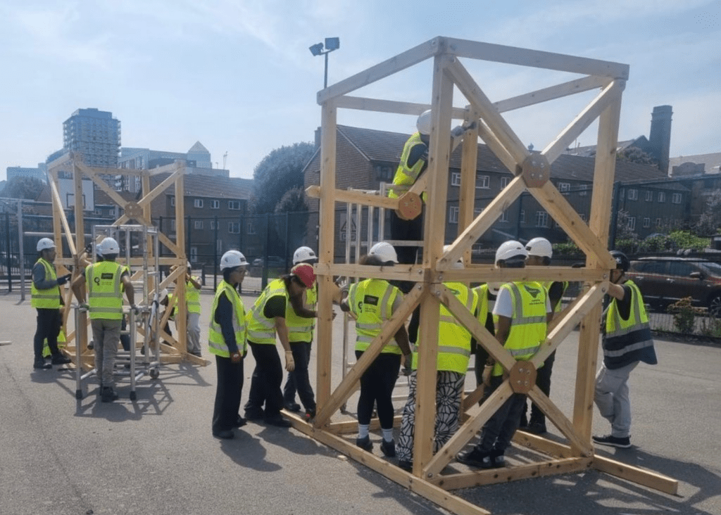 A group of people wearing high-visibility vests and hard hats work together outdoors to assemble large wooden frame structures. The structures are made of timber beams with cross-bracing and metal connectors. Some participants are inside or on the frames, while others stand around assisting. The activity takes place in a paved area with buildings and a fence in the background under a sunny sky.