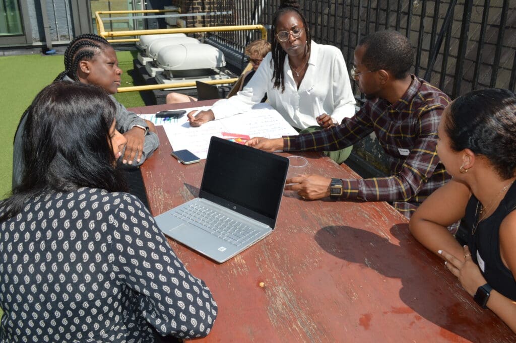 Five people both female and male sitting outside together on bench with one of them having a laptop Infront of them, whilst they speak and discuss amongst themselves.