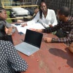 Five people both female and male sitting outside together on bench with one of them having a laptop Infront of them, whilst they speak and discuss amongst themselves.