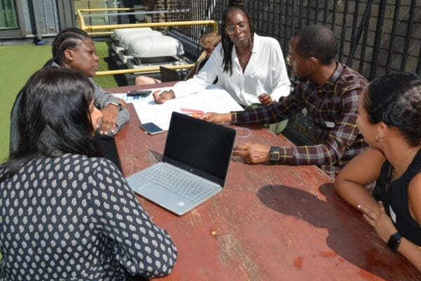 Five people both female and male sitting outside together on bench with one of them having a laptop Infront of them, whilst they speak and discuss amongst themselves.