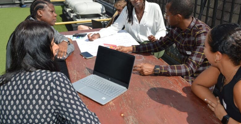 Five people both female and male sitting outside together on bench with one of them having a laptop Infront of them, whilst they speak and discuss amongst themselves.