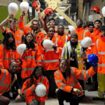 A group of people wearing bright orange high-visibility jackets and safety gear stand together inside a construction site. Most are holding white hard hats in the air, with a few holding red or blue ones. They are posing for a group photo, some kneeling in front and others standing behind. The background shows wooden structures, tools, and construction materials.