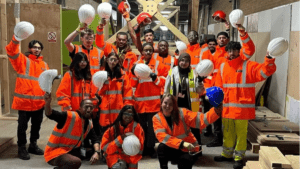 A group of people wearing bright orange high-visibility jackets and safety gear stand together inside a construction site. Most are holding white hard hats in the air, with a few holding red or blue ones. They are posing for a group photo, some kneeling in front and others standing behind. The background shows wooden structures, tools, and construction materials.