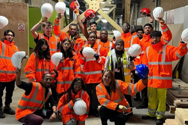 A group of people wearing bright orange high-visibility jackets and safety gear stand together inside a construction site. Most are holding white hard hats in the air, with a few holding red or blue ones. They are posing for a group photo, some kneeling in front and others standing behind. The background shows wooden structures, tools, and construction materials.