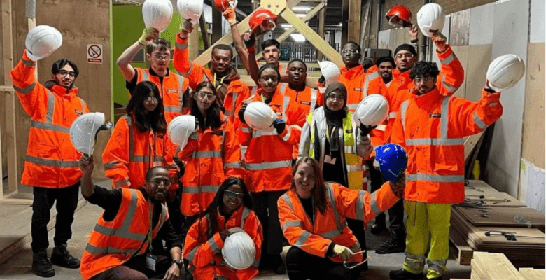 A group of people wearing bright orange high-visibility jackets and safety gear stand together inside a construction site. Most are holding white hard hats in the air, with a few holding red or blue ones. They are posing for a group photo, some kneeling in front and others standing behind. The background shows wooden structures, tools, and construction materials.