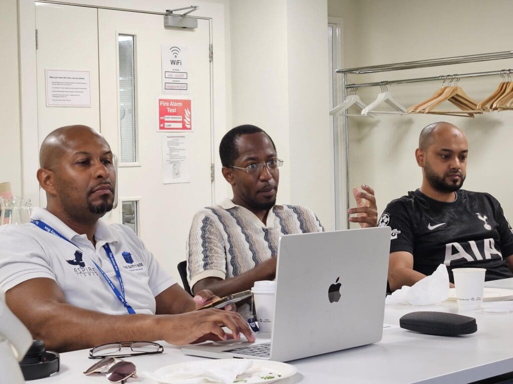 Three men sitting at a conference table during a meeting. The man on the left wears a white polo shirt with lanyards and types on a laptop. The man in the center wears a patterned knit shirt and glasses, gesturing with his hand while looking forward. The man on the right wears a black Tottenham Hotspur football jersey and listens.