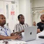 Three men sitting at a conference table during a meeting. The man on the left wears a white polo shirt with lanyards and types on a laptop. The man in the center wears a patterned knit shirt and glasses, gesturing with his hand while looking forward. The man on the right wears a black Tottenham Hotspur football jersey and listens.