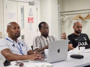 Three men sitting at a conference table during a meeting. The man on the left wears a white polo shirt with lanyards and types on a laptop. The man in the center wears a patterned knit shirt and glasses, gesturing with his hand while looking forward. The man on the right wears a black Tottenham Hotspur football jersey and listens.