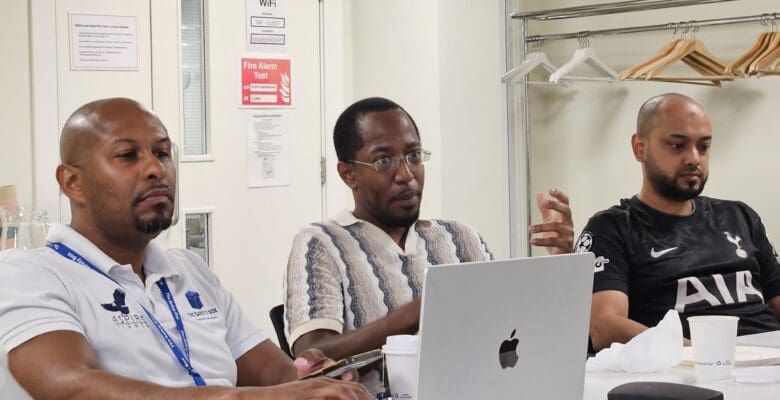 Three men sitting at a conference table during a meeting. The man on the left wears a white polo shirt with lanyards and types on a laptop. The man in the center wears a patterned knit shirt and glasses, gesturing with his hand while looking forward. The man on the right wears a black Tottenham Hotspur football jersey and listens.