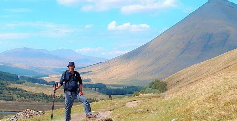 A hiker wearing a bucket hat, backpack, and holding a trekking pole smiles while posing on a dirt trail. The grassy, sloped path winds through a vast valley under a bright blue sky with sparse clouds. In the background, a large, conical green mountain rises steeply on the right side of the frame, while softer, distant mountain ridges fade into the horizon on the left.