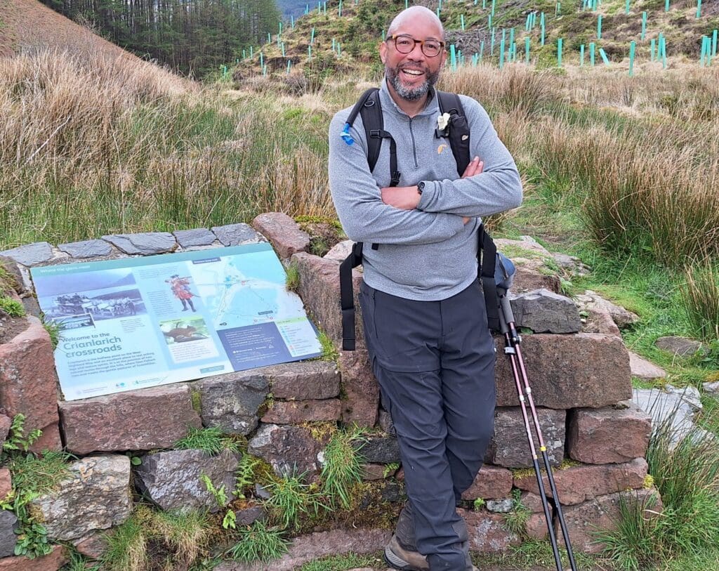 A smiling, middle-aged Black man with glasses, a beard, and a backpack stands outdoors leaning against a stone wall. To his left is an informational sign titled "Welcome to the Crianlarich crossroads." The background shows a grassy hill dotted with young, protected trees.