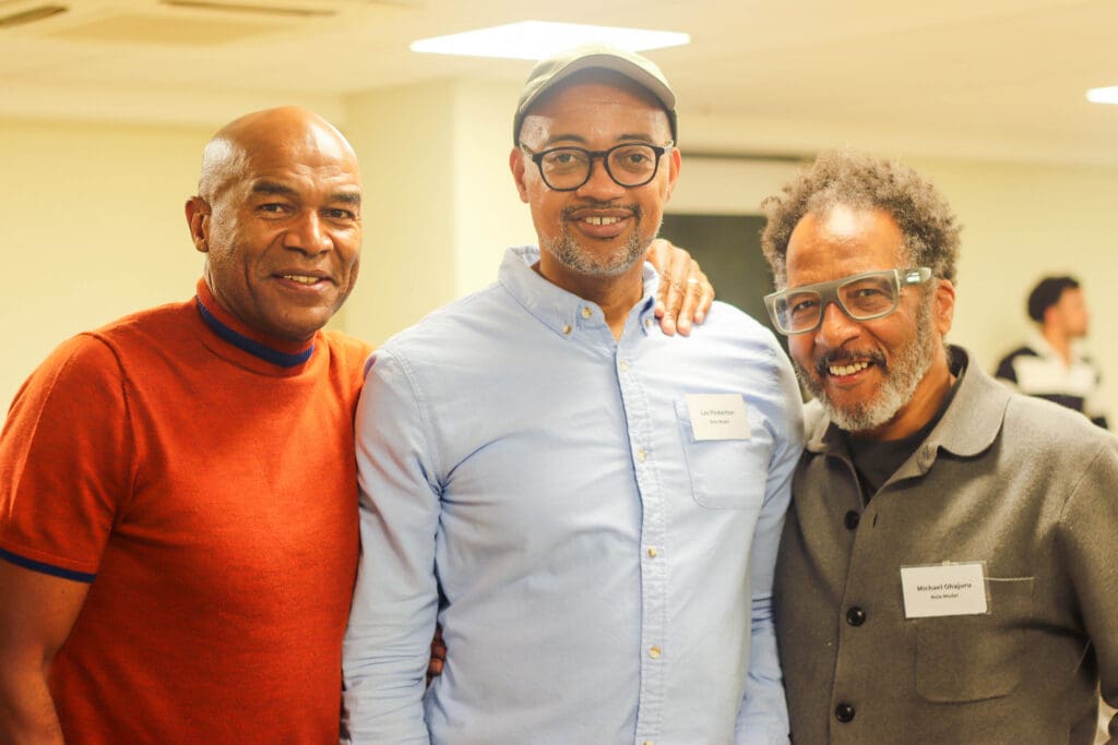 Three men smiling at the camera. the first man is wearing orange t-shirt and has his hand over the second man who is wear a light blue shirt. the Third is wearing a grey cotton jacket.
