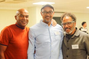 Three men smiling at the camera. the first man is wearing orange t-shirt and has his hand over the second man who is wear a light blue shirt. the Third is wearing a grey cotton jacket.