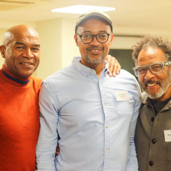 Three men smiling at the camera. the first man is wearing orange t-shirt and has his hand over the second man who is wear a light blue shirt. the Third is wearing a grey cotton jacket.