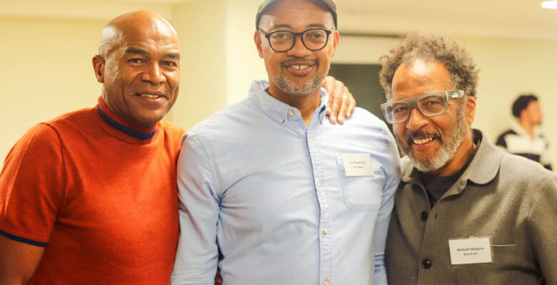 Three men smiling at the camera. the first man is wearing orange t-shirt and has his hand over the second man who is wear a light blue shirt. the Third is wearing a grey cotton jacket.